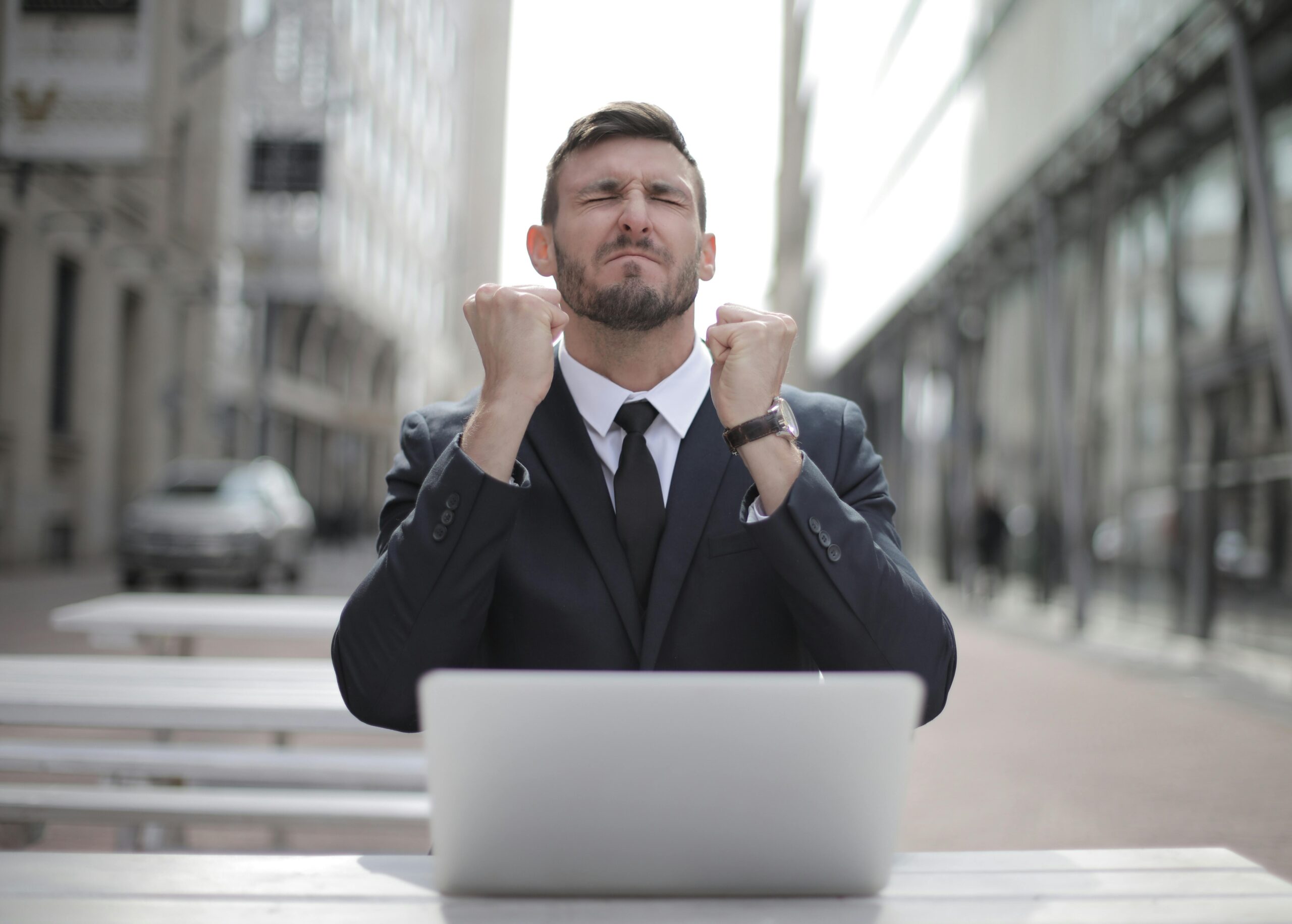 A man in a suit looking joyful in front of a laptop while sitting on a bench.