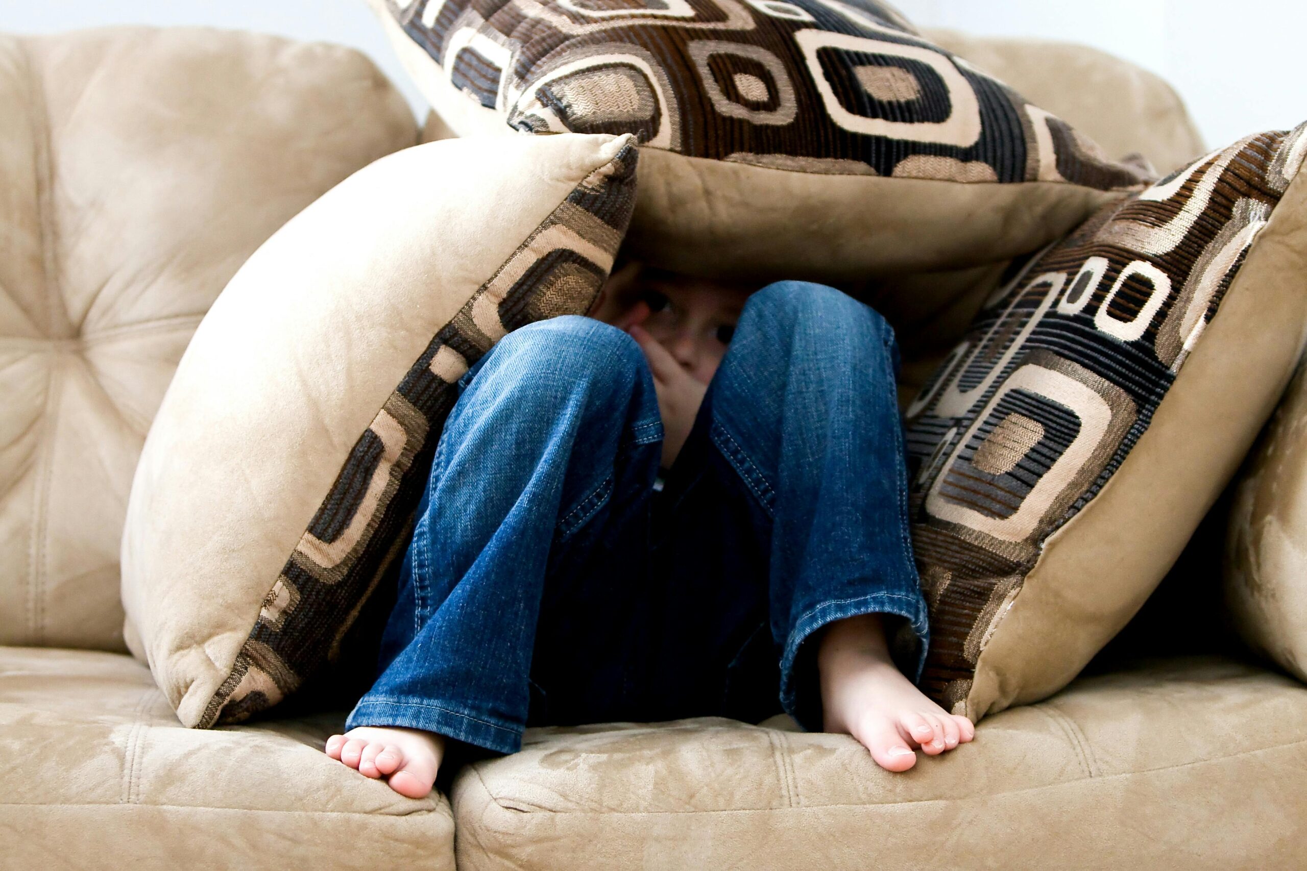 A jean wearing bare foot kid is laying on a couch covered in cushions.